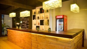 Un espace d’accueil avec un comptoir en bois, des étagères remplies de bouteilles d’alcool, un réfrigérateur Coca-Cola et des lampes suspendues.