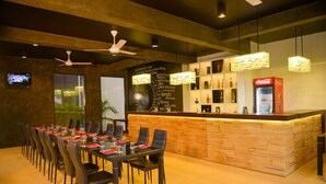 Un restaurant avec une longue table dressée pour le dîner, un coin bar avec un réfrigérateur rempli de Coca-Cola et un mur où est affichée la carte des vins.