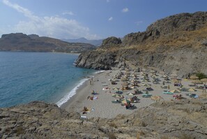 A beach with rocky cliffs, a sandy shore, and umbrellas. - Ammoudi Hotel (Agios Vasileios)