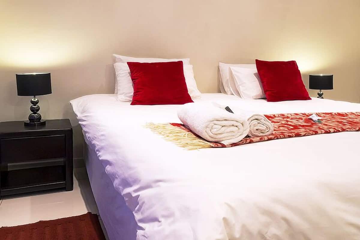 A neatly made bed with white and red pillows, a rolled towel, and a red and white patterned blanket. There are two black bedside tables with lamps.