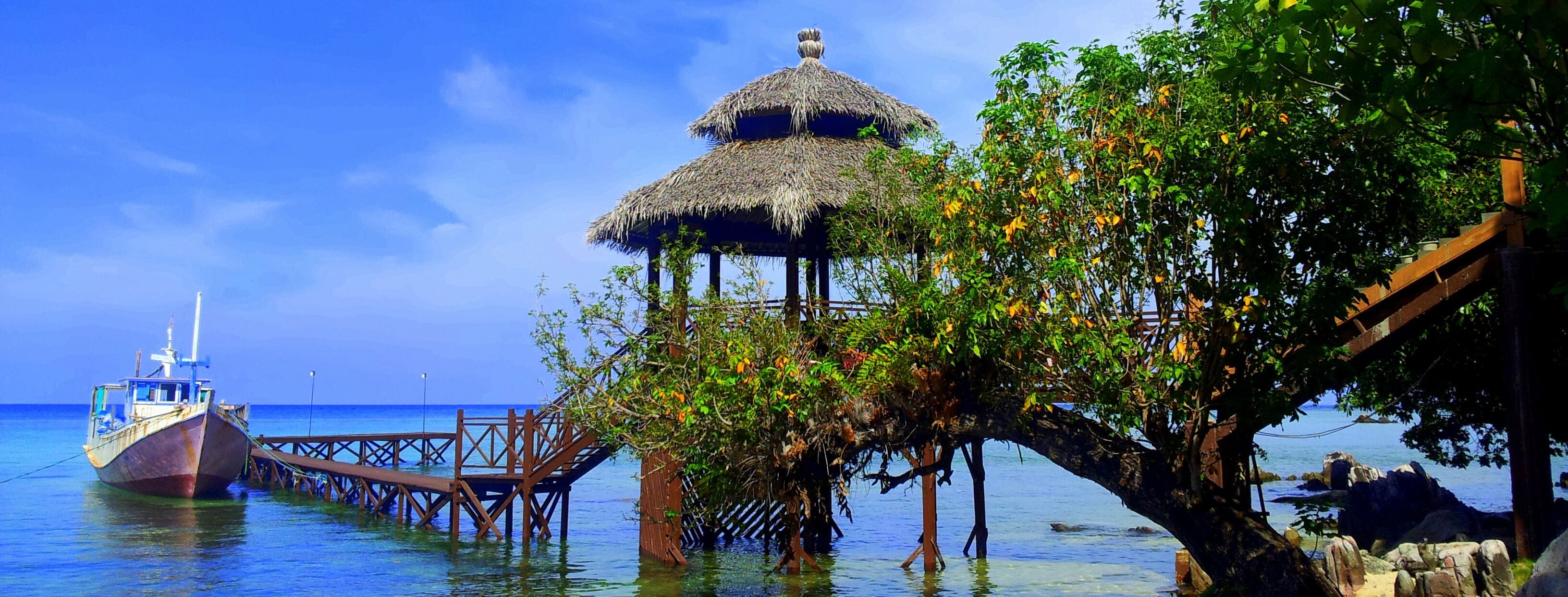 A wooden pier extending into the water with a thatched-roof structure and a boat docked at the end.