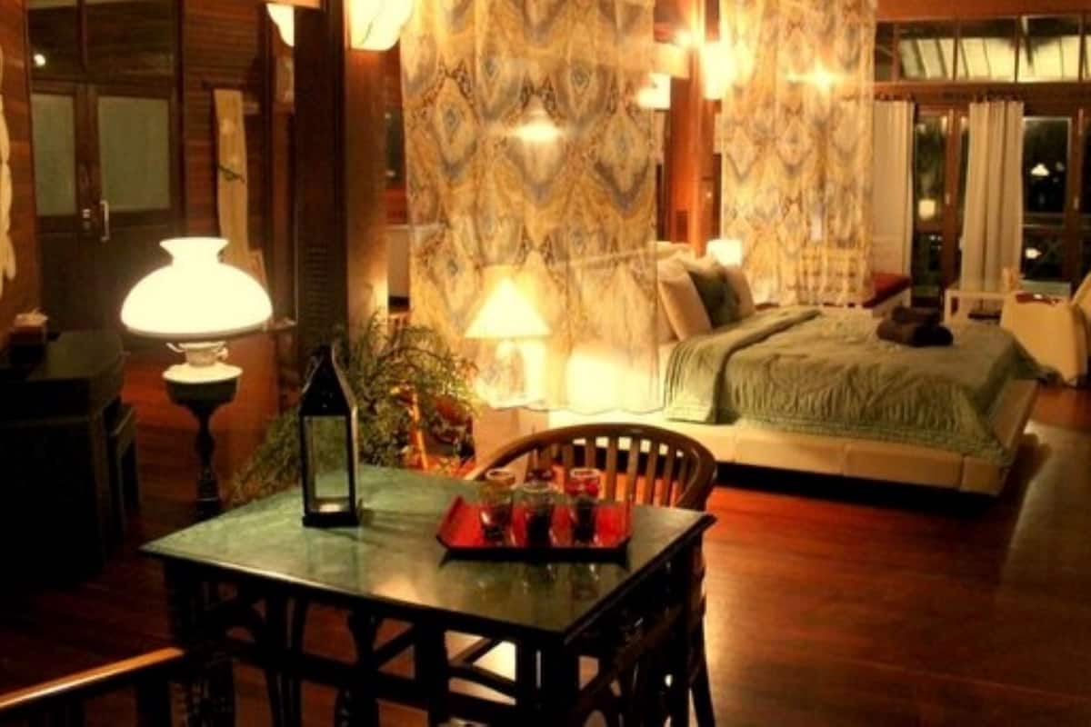 Una habitación de hotel acogedora con piso de madera, una cama con manta verde, una mesa de comedor con lámpara y una silla.