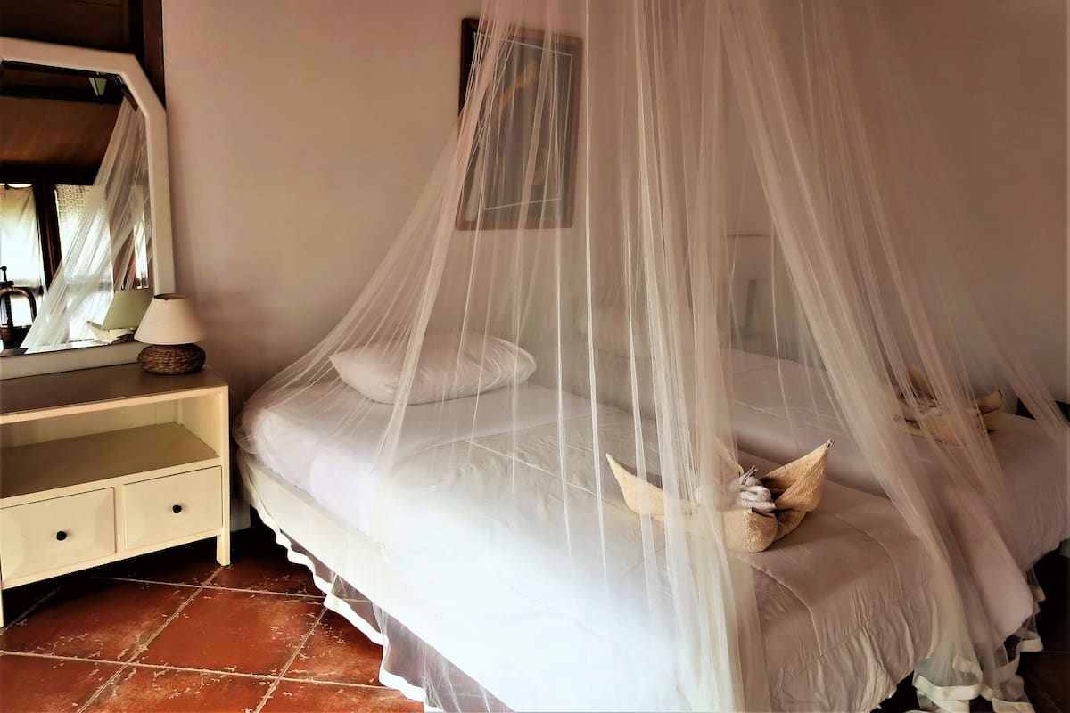 Un dormitorio con cama bajo dosel, una mesita de noche con lámpara y suelo de baldosas.