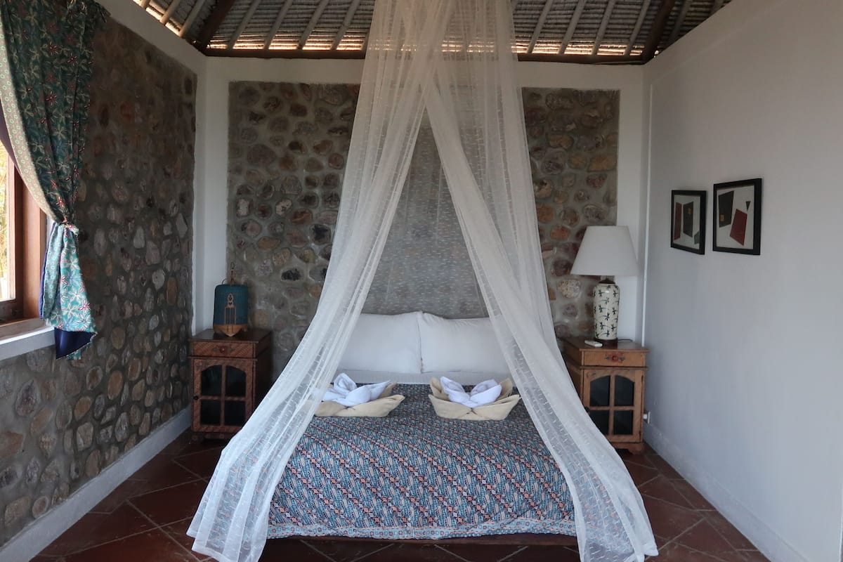 Un dormitorio con pared de piedra, una cama con colcha estampada, una mesita de noche con lámpara y una ventana con cortinas.