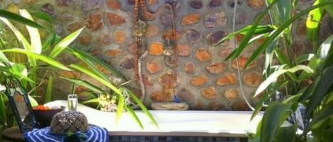Un espace patio agrémenté d’un mur en mosaïque, d’une petite table sur laquelle repose un chapeau, et de diverses plantes.
