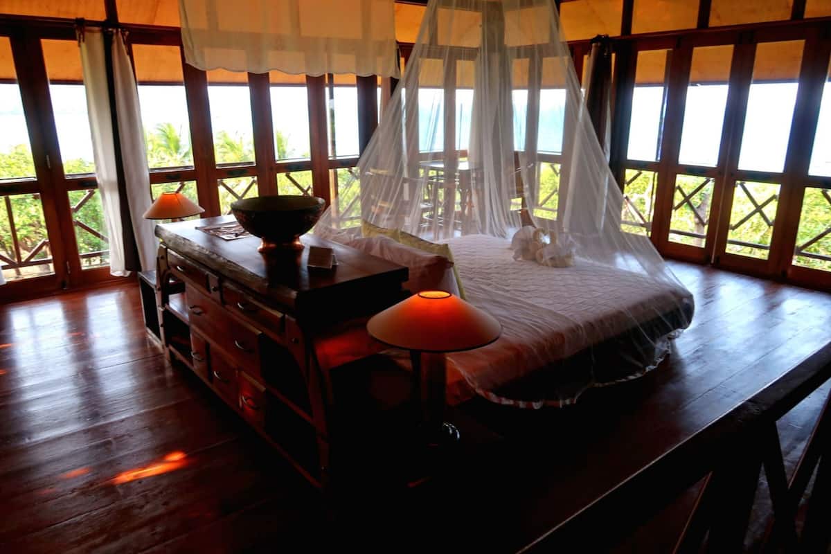 Habitación con cama bajo una mosquitera, muebles de madera y ventanas amplias con vistas a la vegetación.