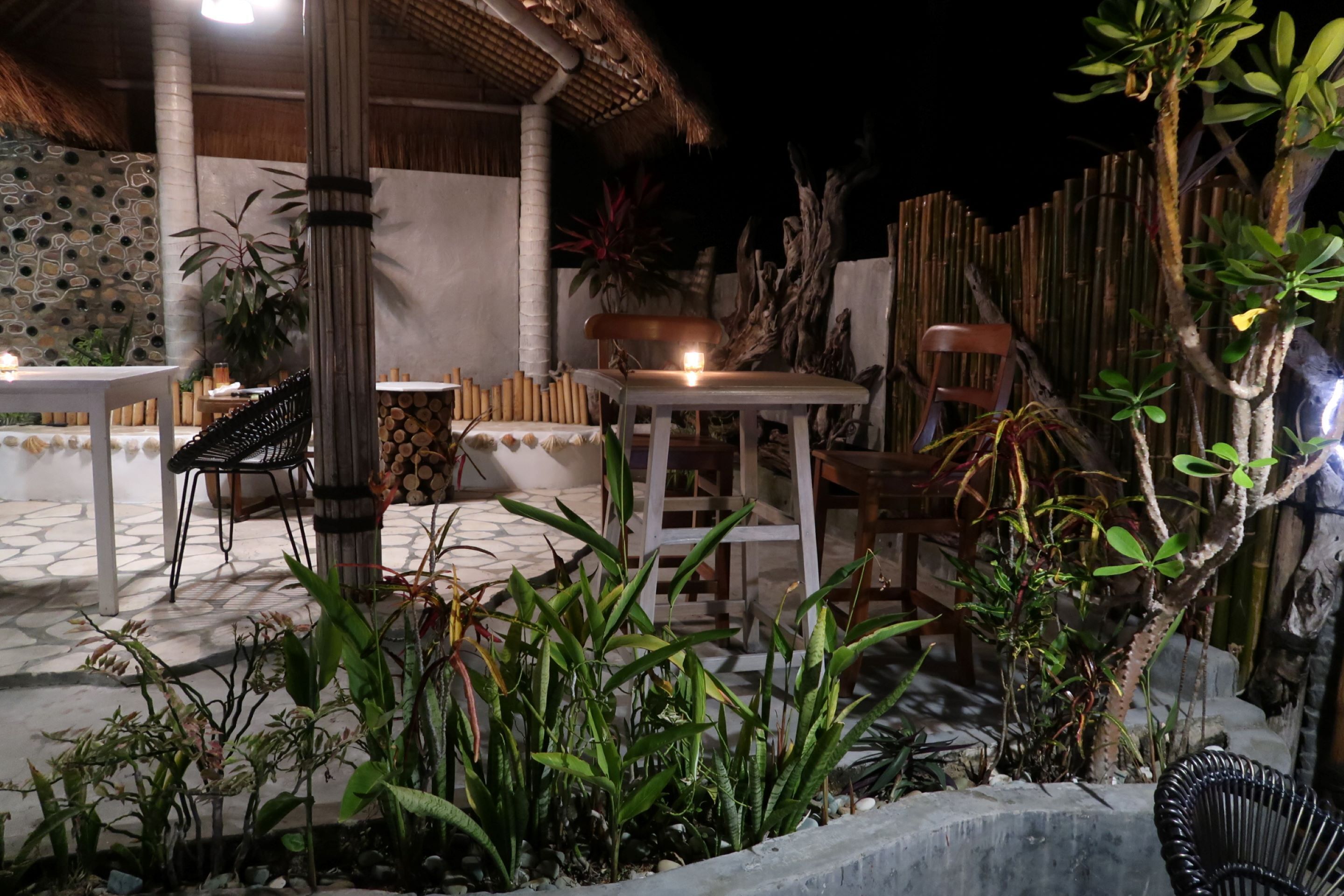 Un espace extérieur aménagé en terrasse, avec des tables et des chaises, entouré de plantes et surmonté d’une structure à toit de chaume.