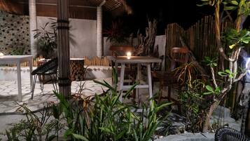 Un espace extérieur aménagé en terrasse, avec des tables et des chaises, entouré de plantes et surmonté d’une structure à toit de chaume.