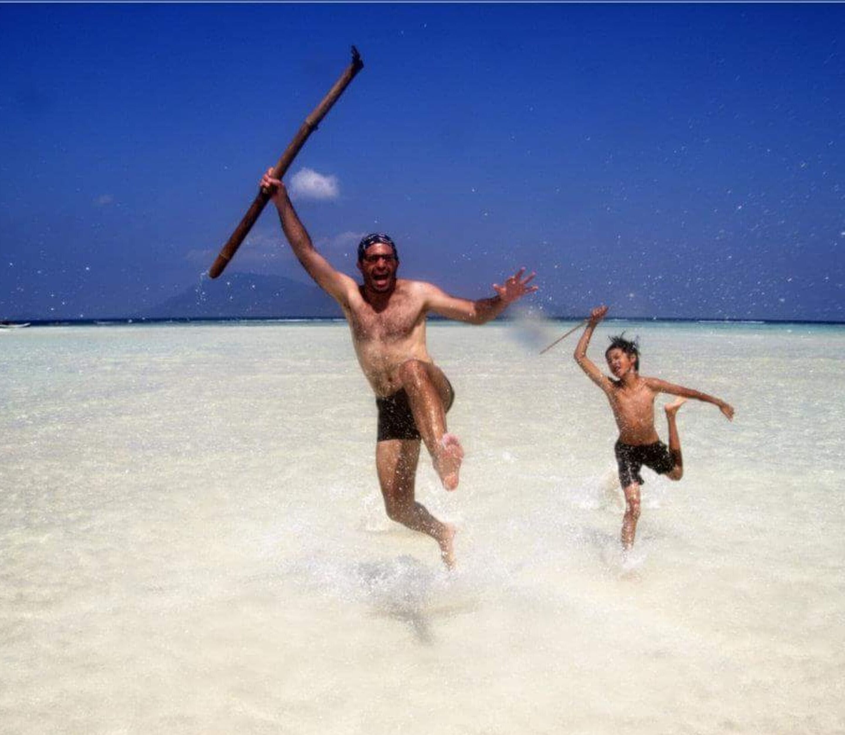 Two people running in shallow water with a clear sky and a distant mountain in the background.