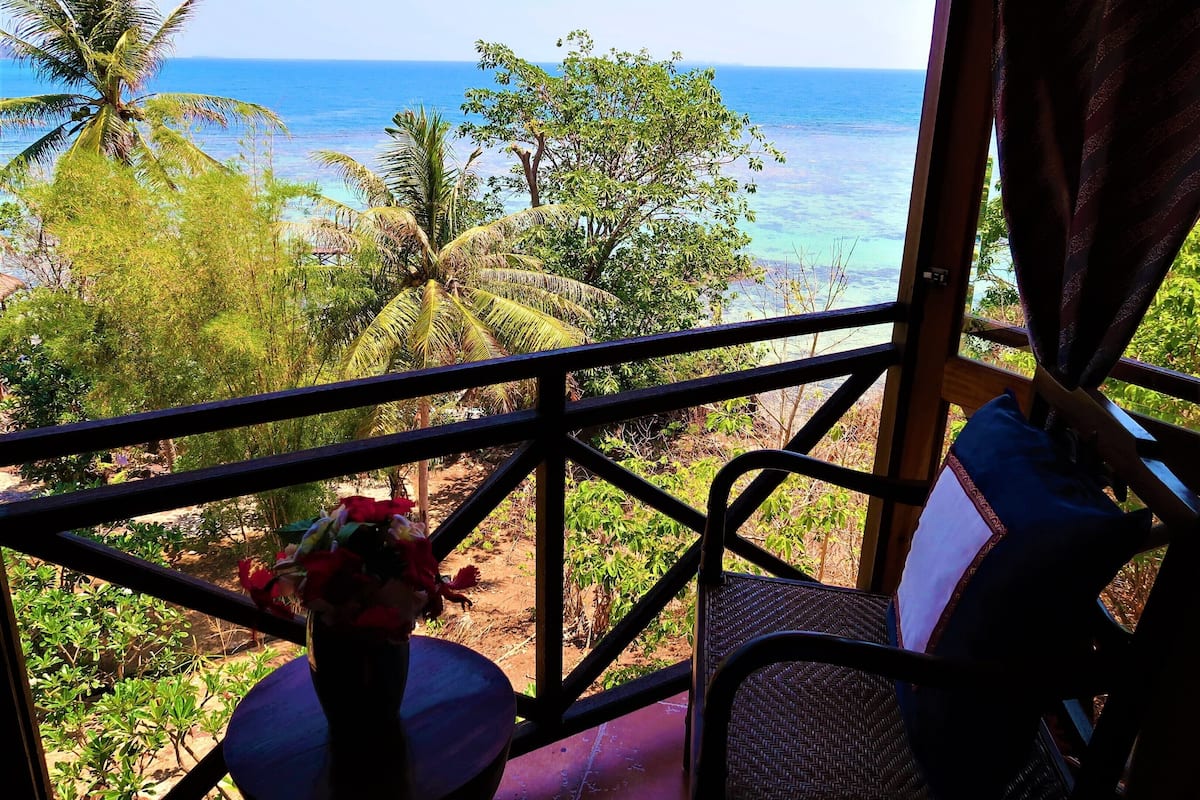 Un balcón con vista al océano, una silla y una mesa con un jarrón de flores.