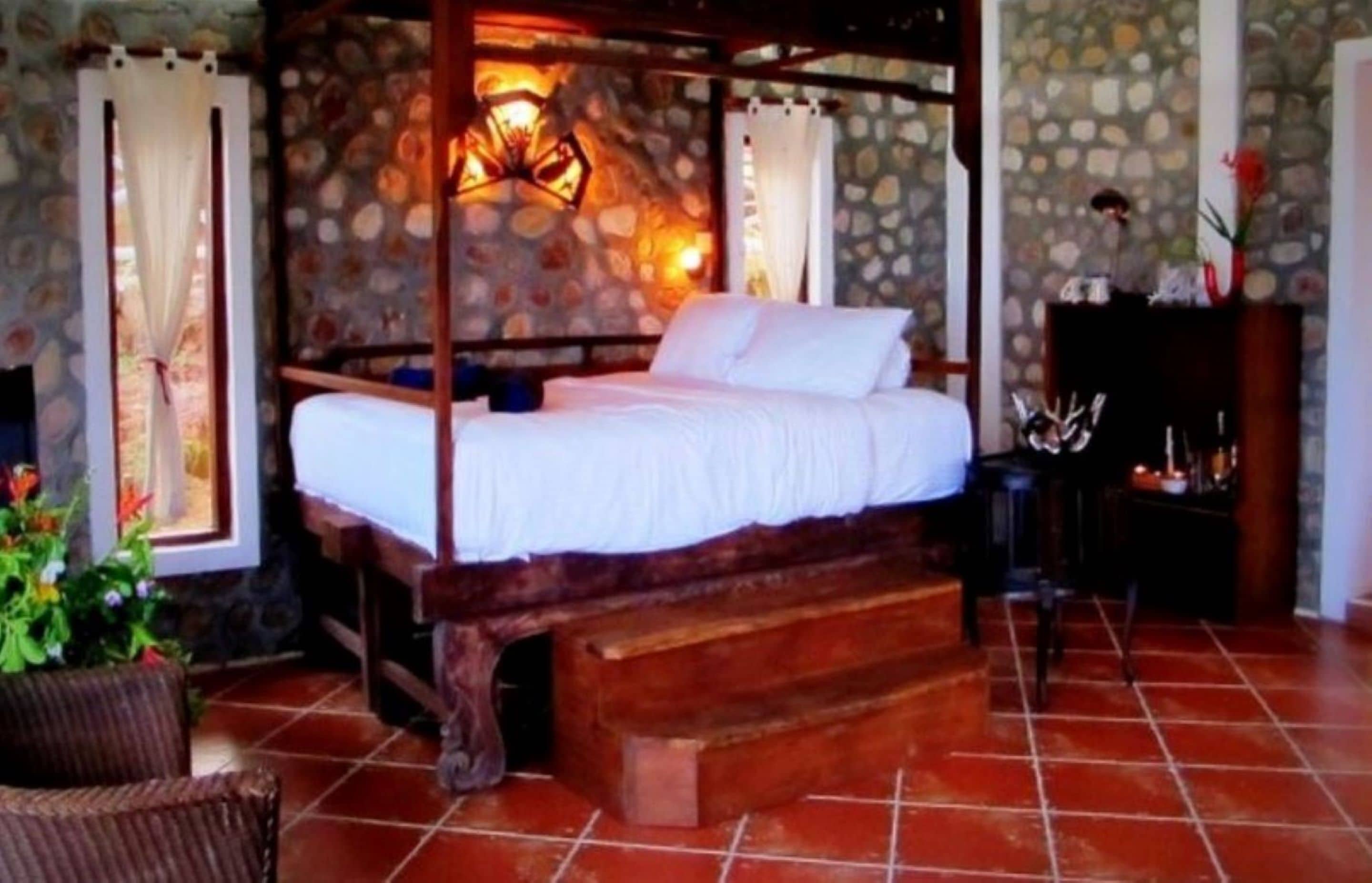A four-poster bed with white linens, a stone wall backdrop, and a rustic wooden bench.