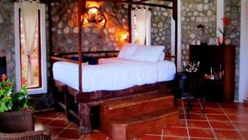 A four-poster bed with white linens, a stone wall backdrop, and a rustic wooden bench.