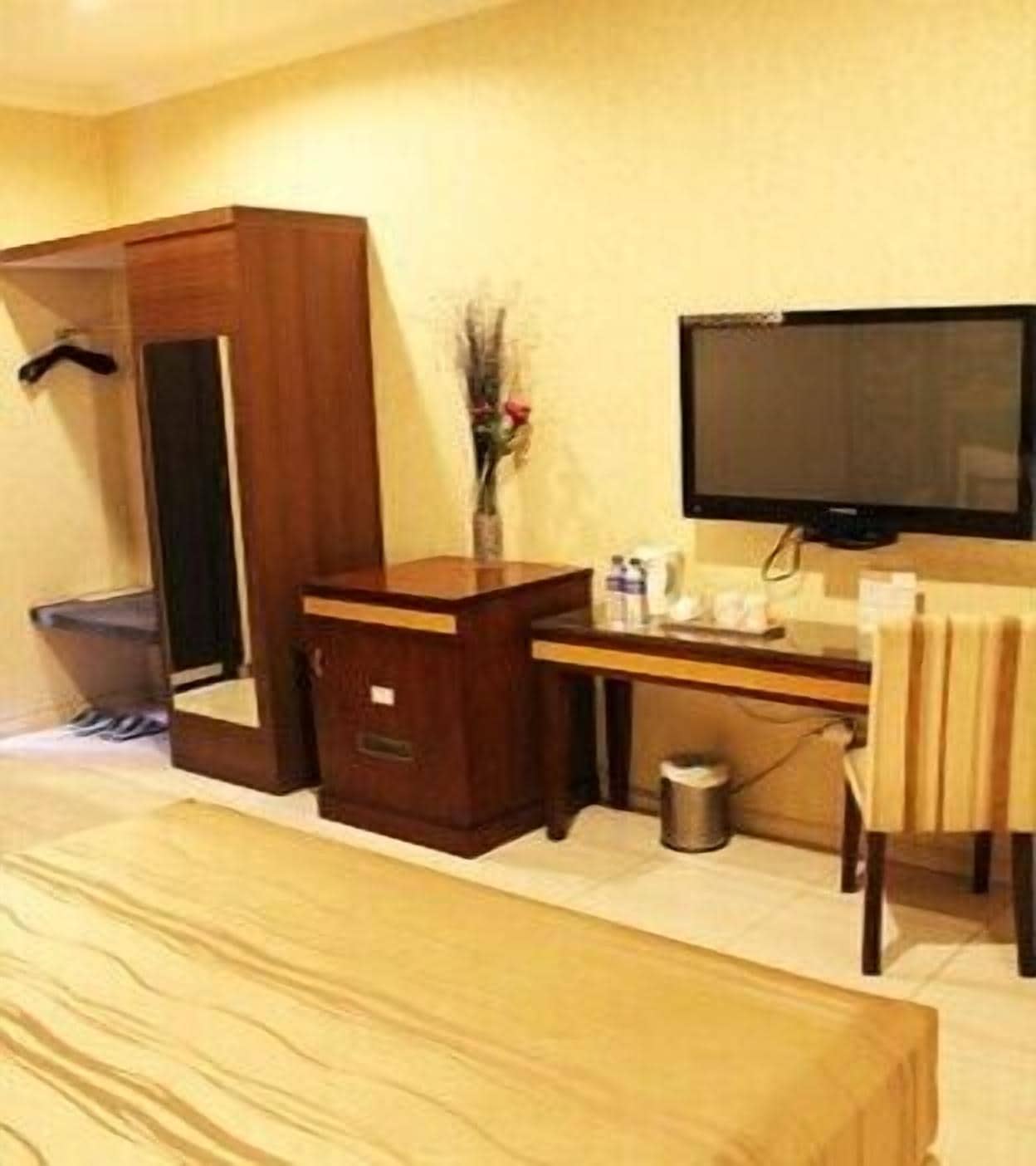 A hotel room with a bed, a wooden wardrobe, a desk with a chair, a television, and a small trash bin.