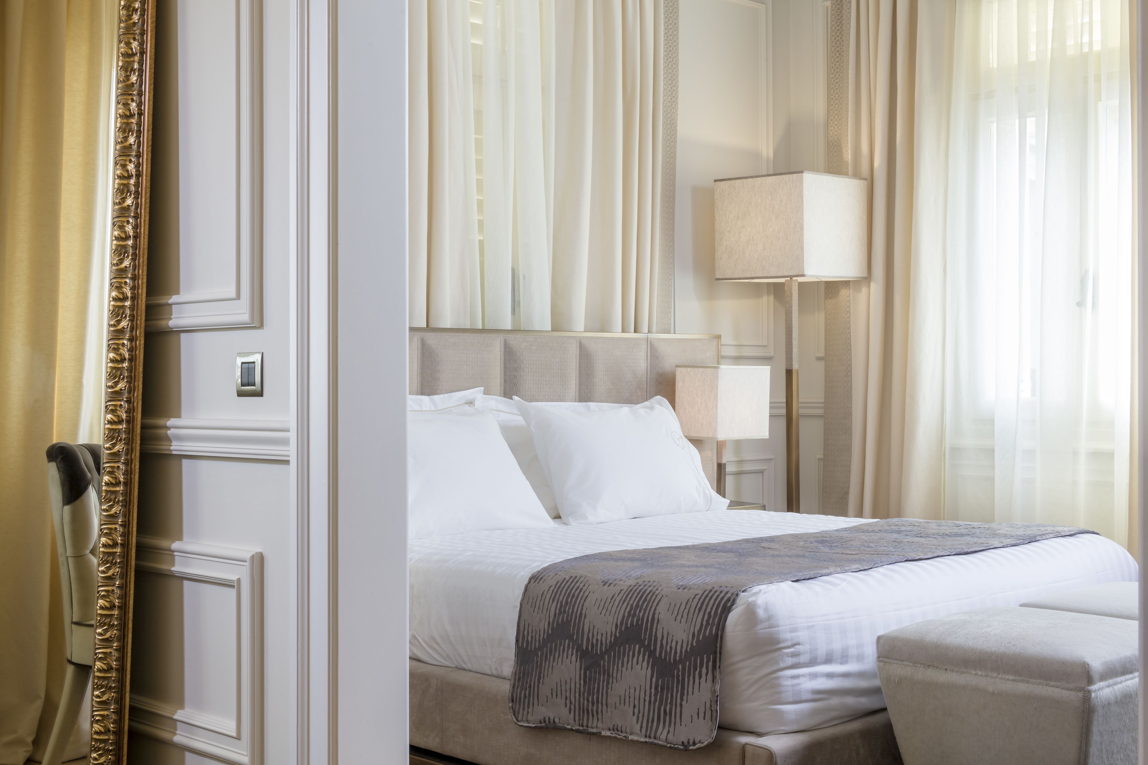 A hotel room with a large bed, a bedside table, a lamp, and a view of the outside through sheer curtains.