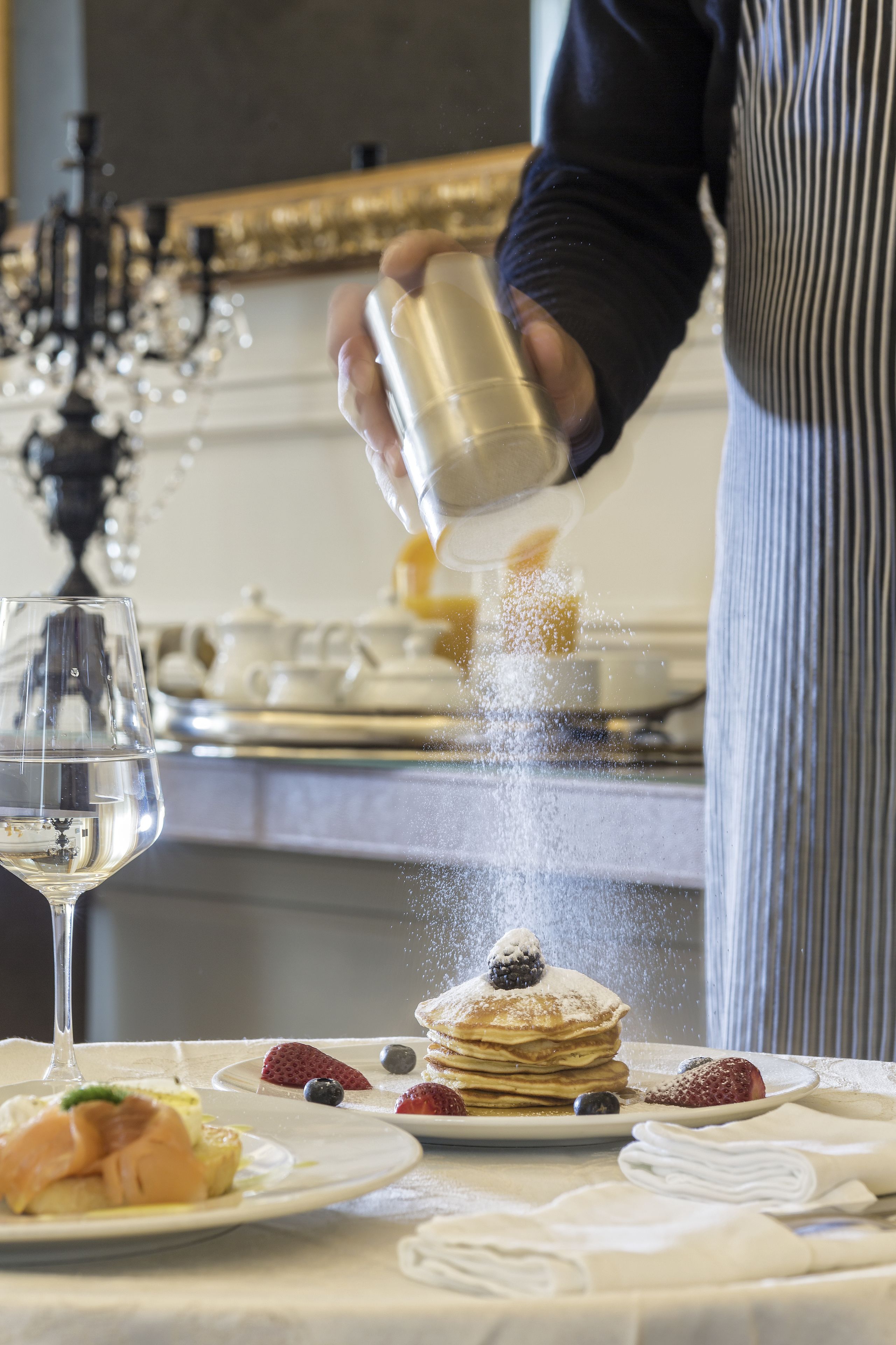 A person sprinkling powdered sugar over a stack of pancakes on a white tablecloth.