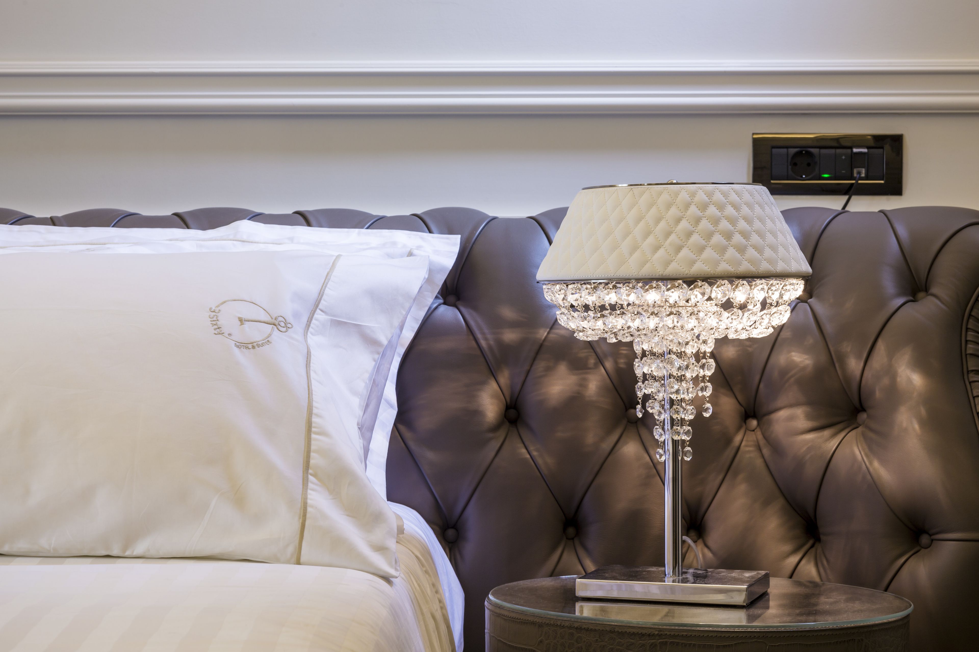 Una cama con edredón blanco y almohada decorativa, cabecera tapizada de cuero, mesita de noche con lámpara de cristal y enchufe en la pared.