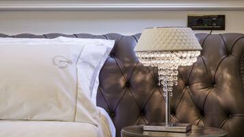 Una cama con edredón blanco y almohada decorativa, cabecera tapizada de cuero, mesita de noche con lámpara de cristal y enchufe en la pared.