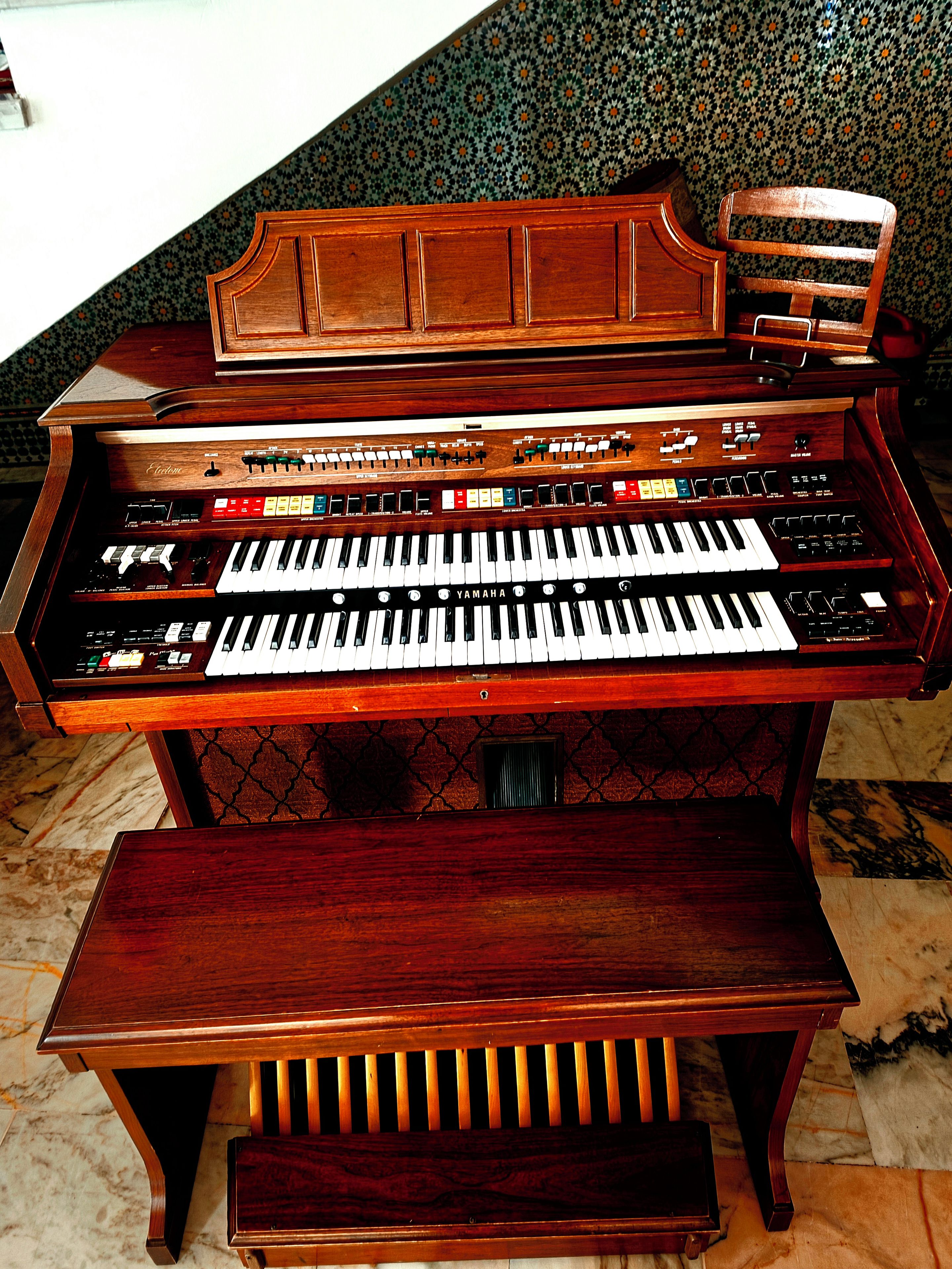 A grand piano with a wooden finish and a bench, placed in a room with patterned wallpaper.