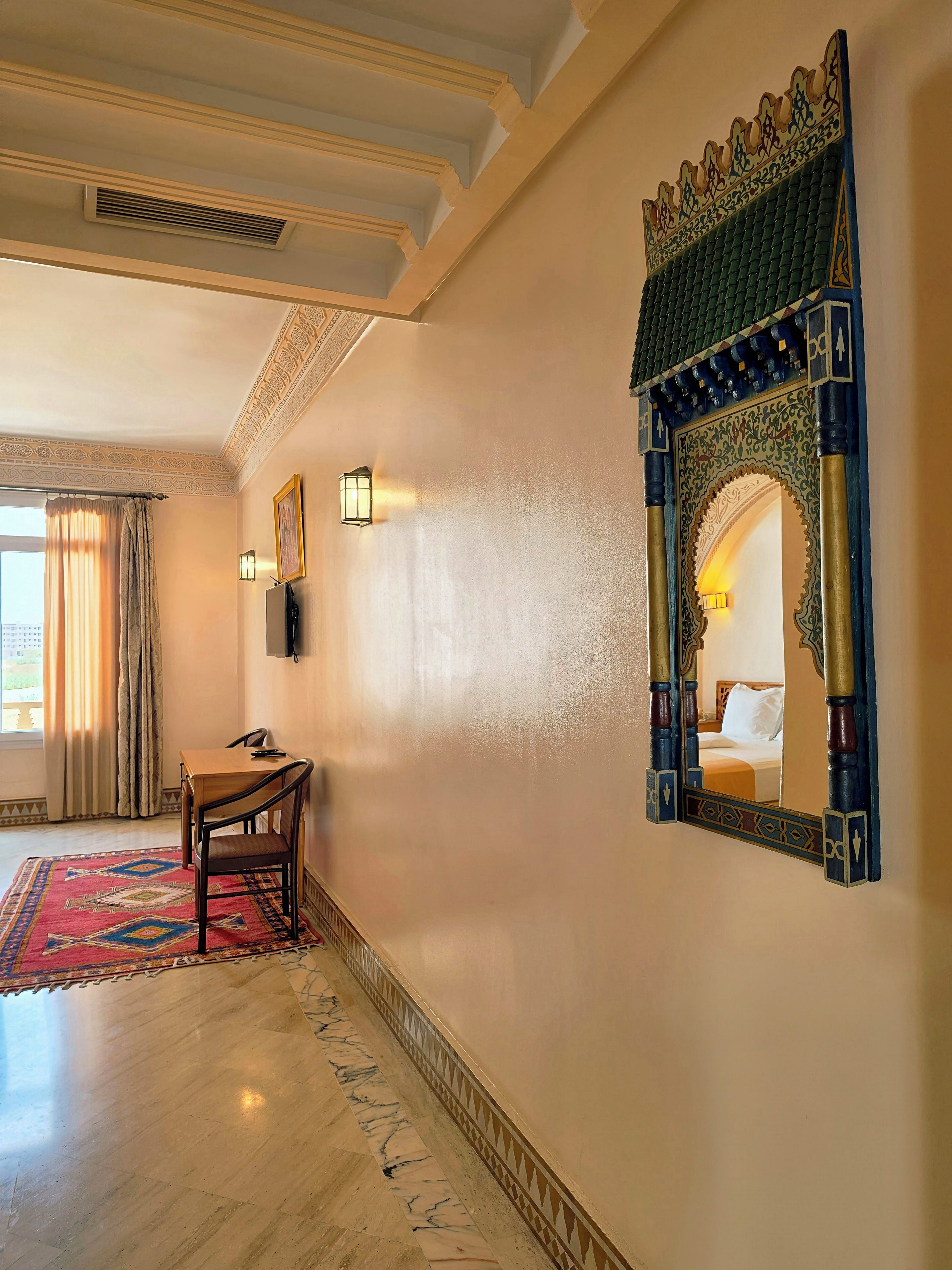 Habitación de hotel con cama, escritorio, silla, espejo decorativo y alfombra estampada.