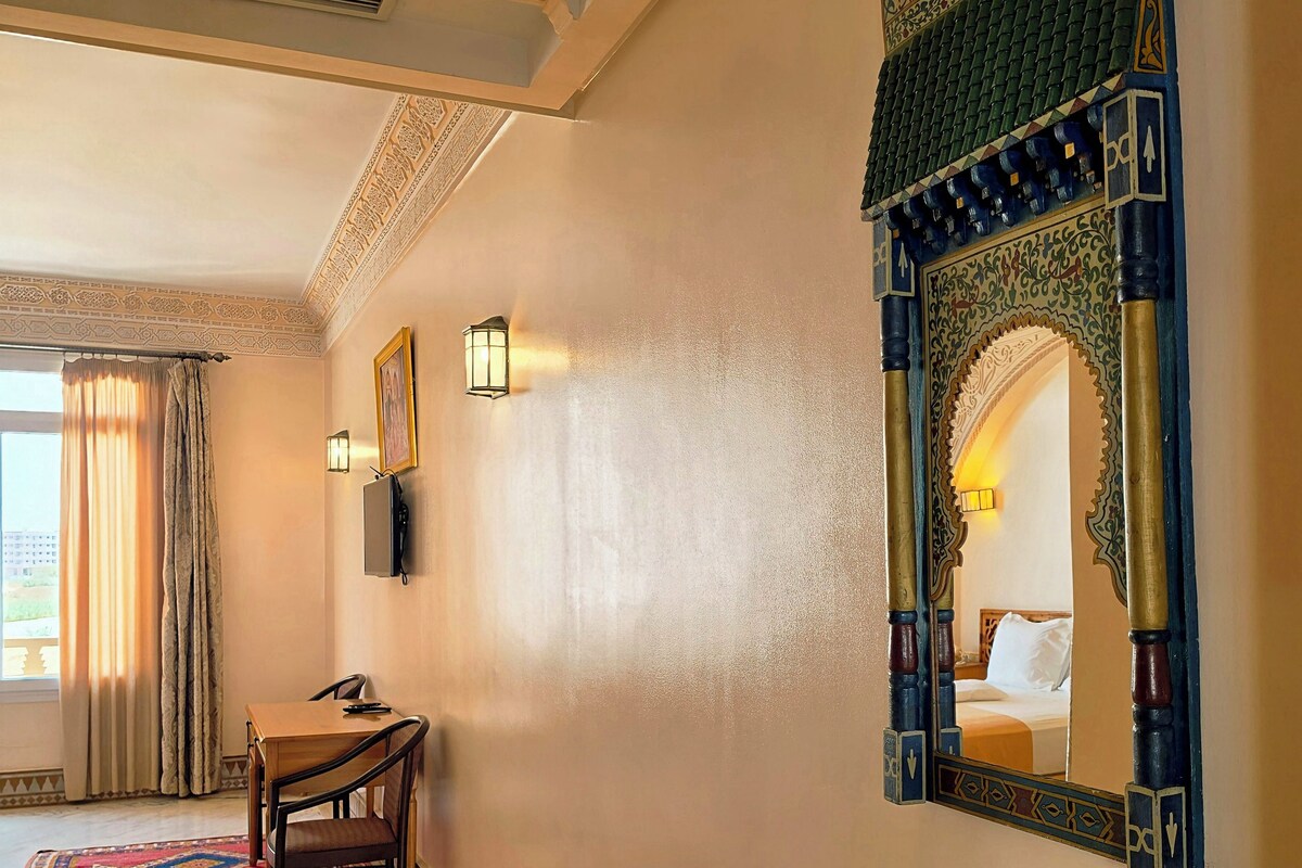 Une chambre d’hôtel comprenant un lit, un bureau, une chaise, un miroir décoratif et un tapis à motifs.