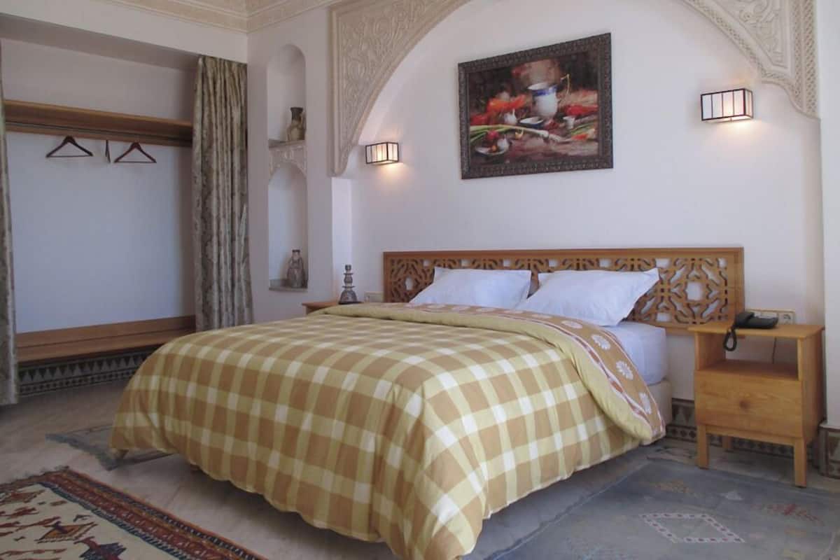 A bedroom with a checkered bedspread, a wooden headboard, a nightstand, a wall-mounted lamp, and a painting above the bed.