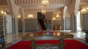 A grand lobby with a red carpet, ornate columns, a chandelier, and a staircase leading to upper floors.