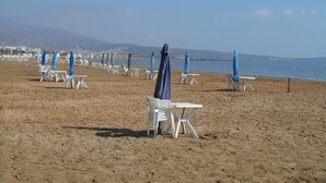A beach with rows of white plastic chairs and tables, some with blue umbrellas, under a clear sky.