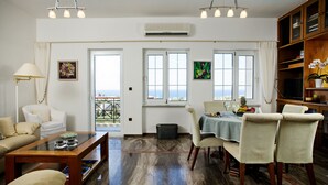 A spacious living room with a sofa, coffee table, and dining area. There are large windows with curtains, an air conditioning unit, and a view of the outdoors. - Star Daratsos (Chania)