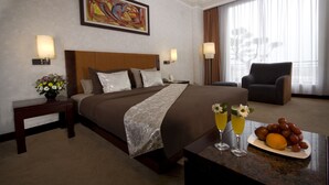 A hotel room with a large bed, a chair, a dining table with fruit and drinks, and a painting on the wall. - Grand Wahid Hotel Salatiga (Salatiga)