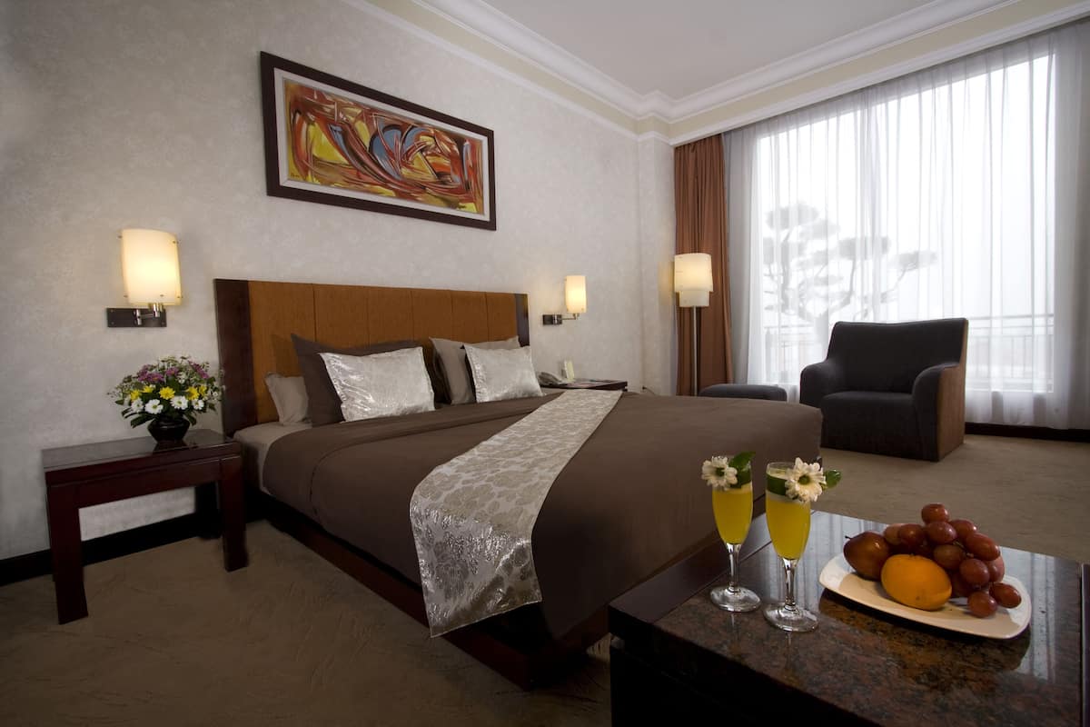 Ein Hotelzimmer mit einem großen Bett, einem Sessel, einem Esstisch mit Obst und Getränken sowie einem Bild an der Wand.