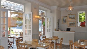 A dining area with wooden tables and chairs, large windows, and a chandelier. - Zefiros Traditional Hotel (Corfu)