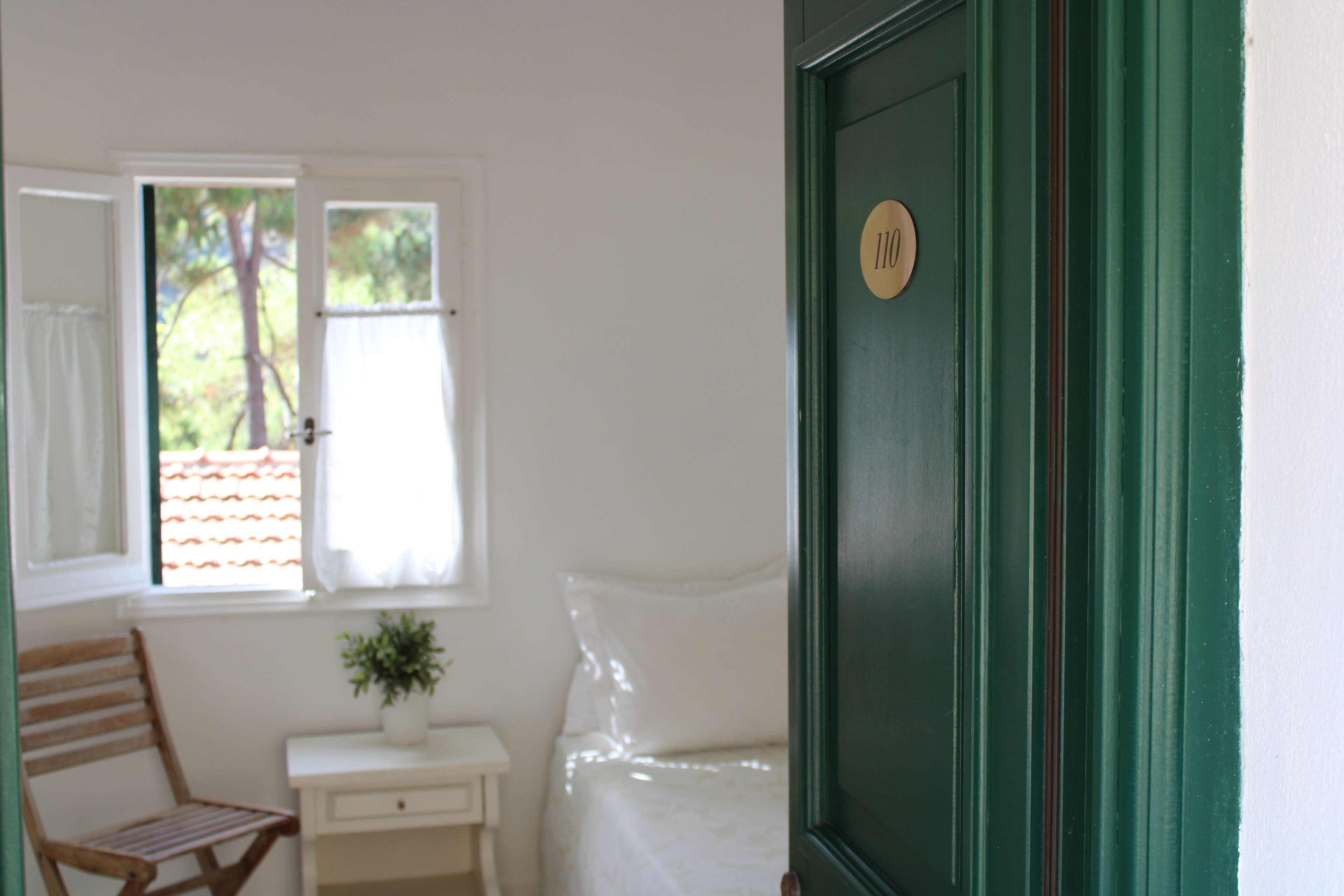 A room with a bed, a chair, a small table with a plant, and a door with a number plate.