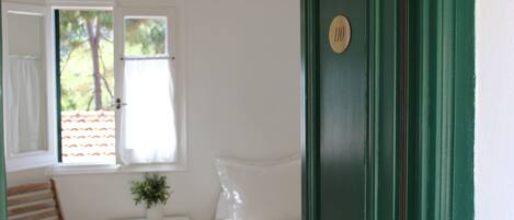 A room with a bed, a chair, a small table with a plant, and a door with a number plate.