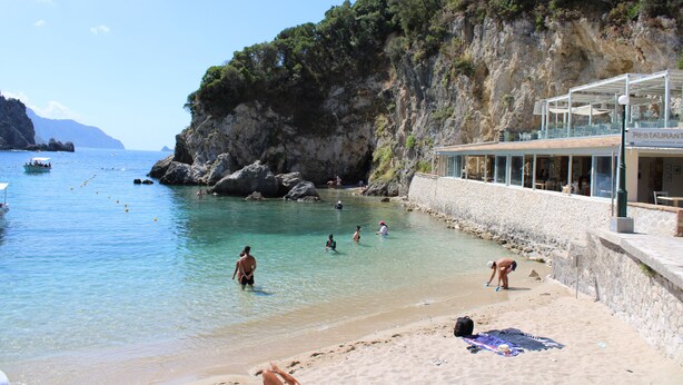A coastal beach with clear waters, rocky cliffs, and a restaurant with outdoor seating. - Zefiros Traditional Hotel (Corfu)