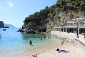 Uma praia costeira com águas claras, falésias rochosas e um restaurante com área externa.