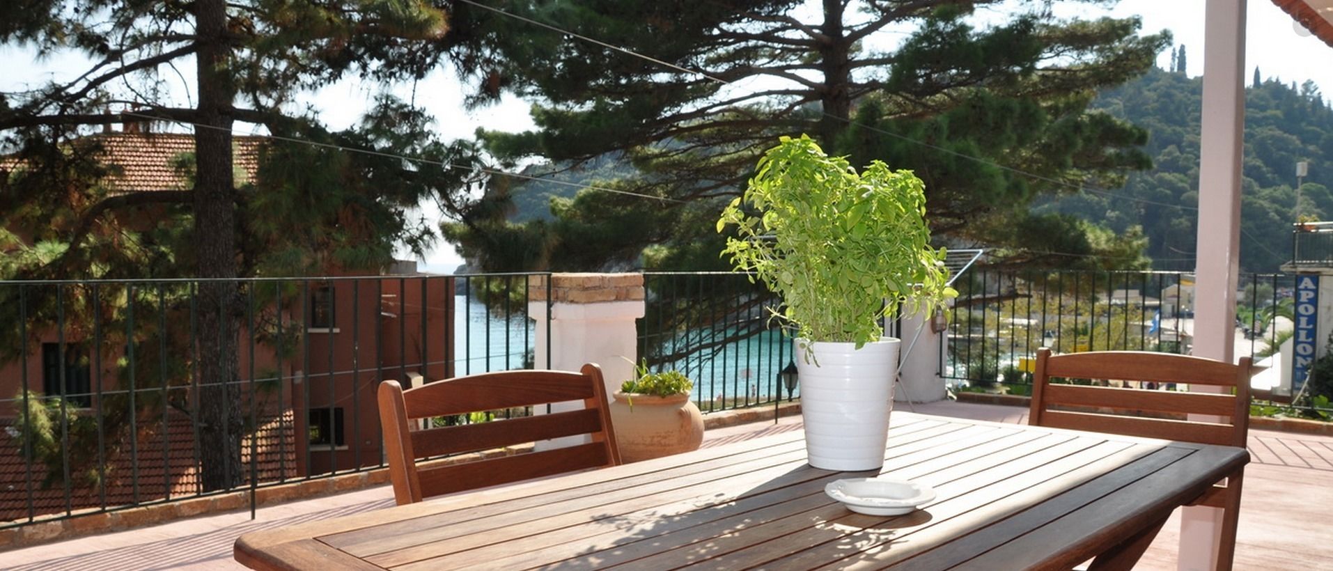 A wooden table with two chairs on a balcony, a potted plant, and a view of the sea and trees.