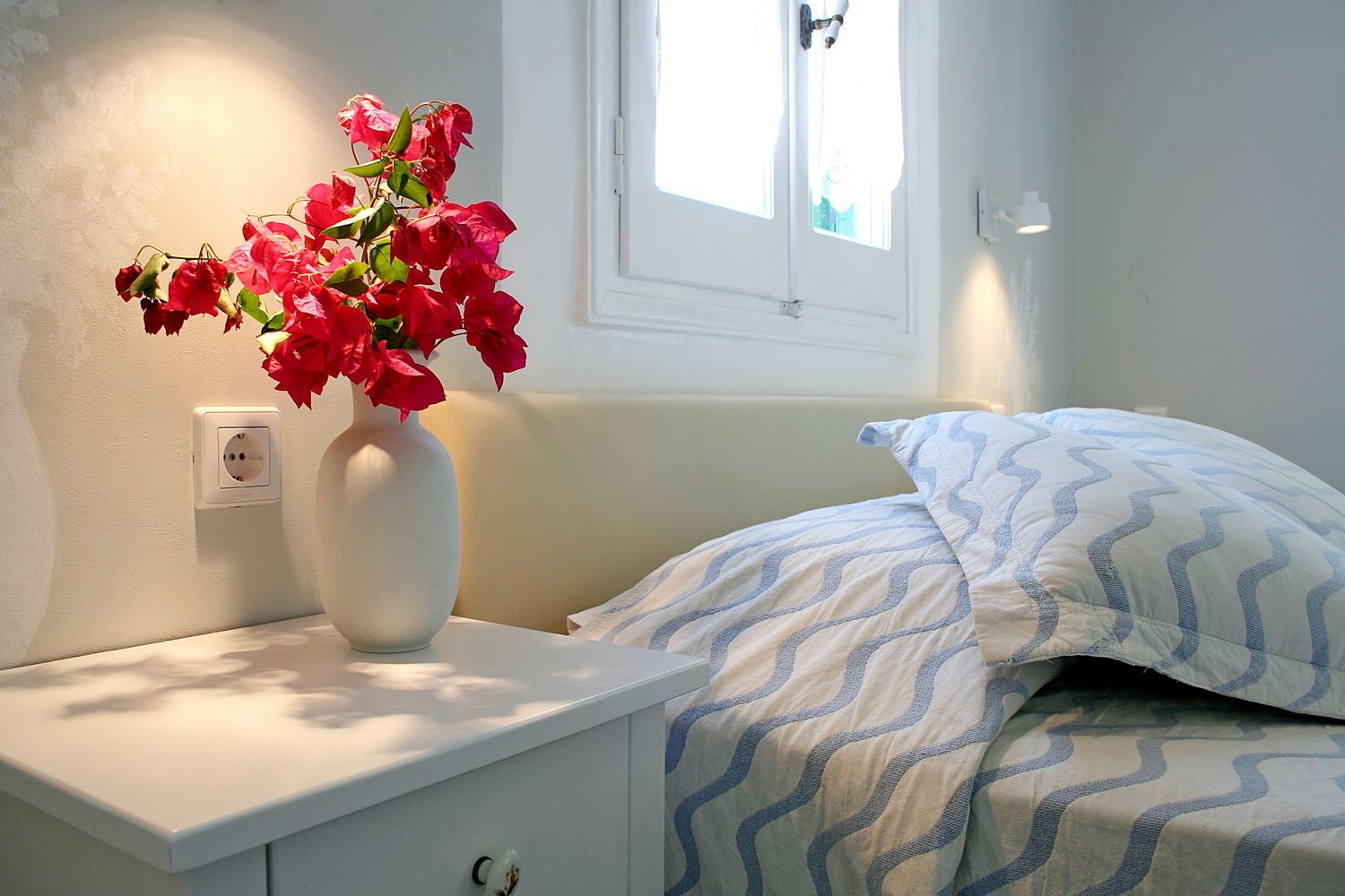 A bedroom with a white bedside table, a vase with red flowers, a patterned pillow, and a window with daylight.