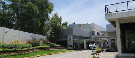 A modern hotel entrance with a paved driveway, well-maintained landscaping, and a signboard advertising room types.