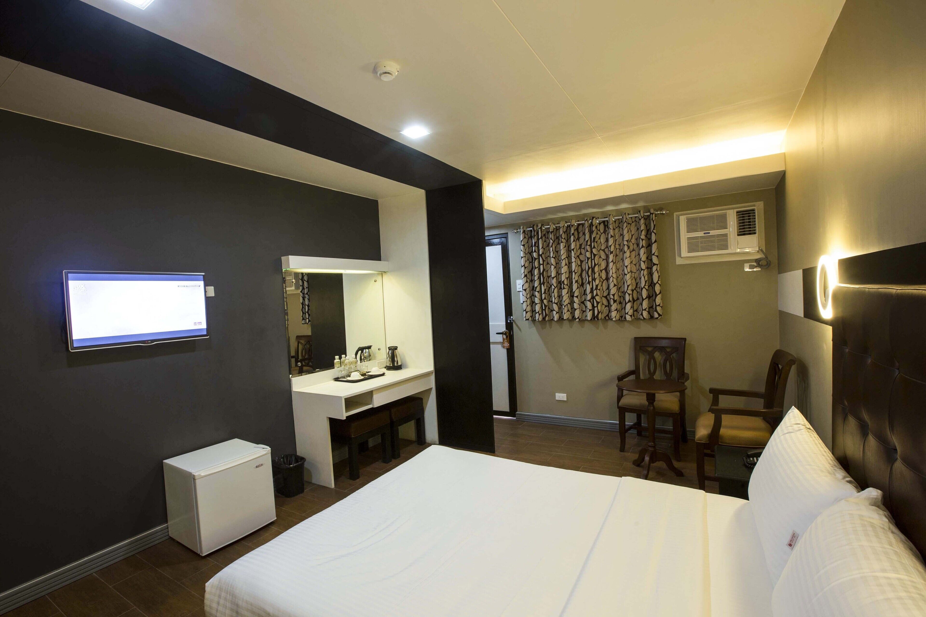 A hotel room with a bed, a desk with a mirror, a small refrigerator, a chair, and a television mounted on the wall.