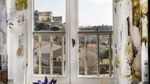 Quarto com uma grande janela com vista para um edifício histórico. Há uma cesta de vime e um pequeno vaso com raminhos na janela.