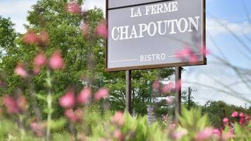 Une enseigne indiquant « Hostellerie La Ferme Chapouton Bistro », avec une vue dégagée sur la verdure environnante et une serre dont on aperçoit une partie.