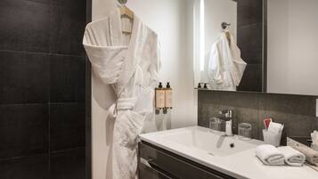 Un baño moderno con un espejo grande, baldosas oscuras, una bañera blanca y un lavabo con artículos de aseo.