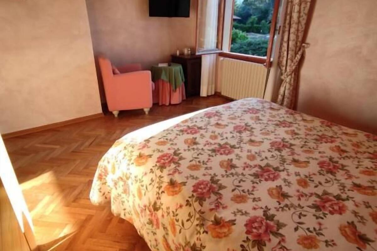 A bedroom with a floral bedspread, a pink chair, a small table, a TV mounted on the wall, and a window with curtains.