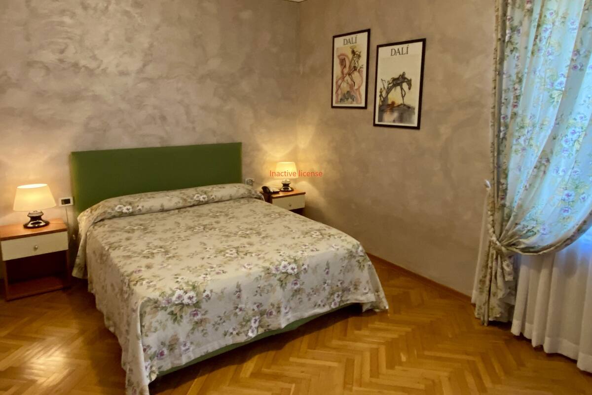 A bedroom with a green headboard, floral bedspread, wooden flooring, a nightstand with a lamp, and two framed pictures on the wall.