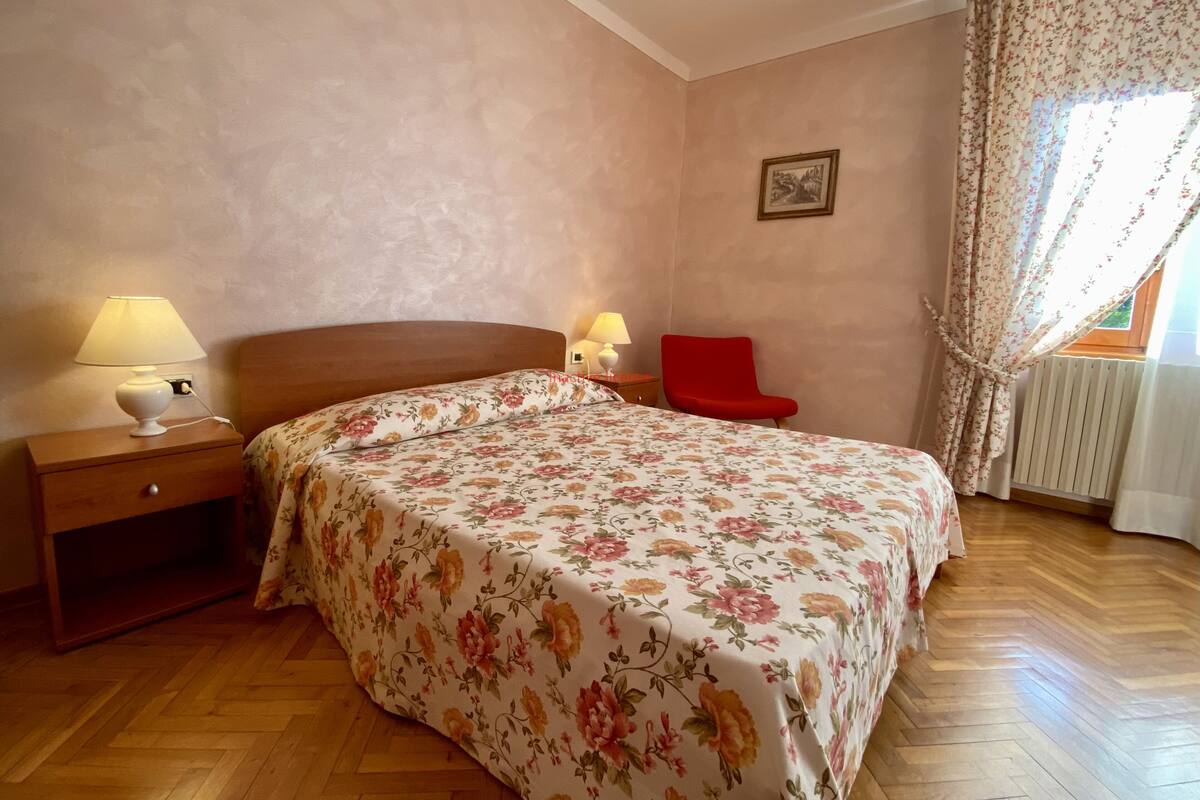 A bedroom with a wooden bed, two bedside lamps, a red chair, a radiator, and a window with floral curtains.