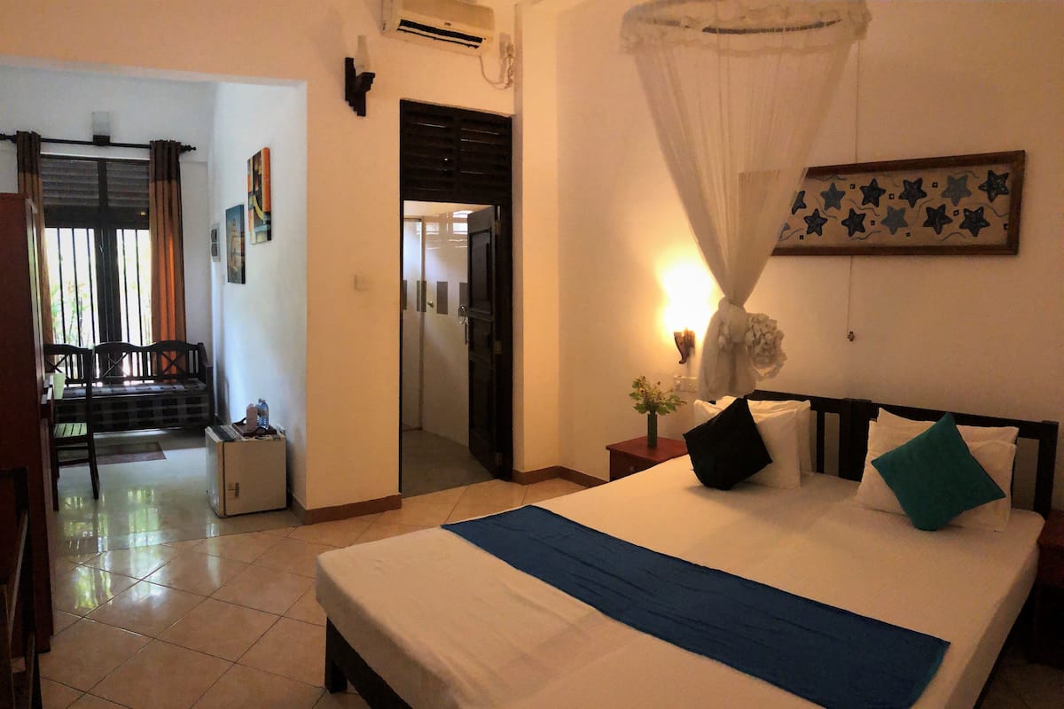A hotel room with a bed, a bedside table, a nightstand, a window with curtains, a door, and a view of the outside balcony.