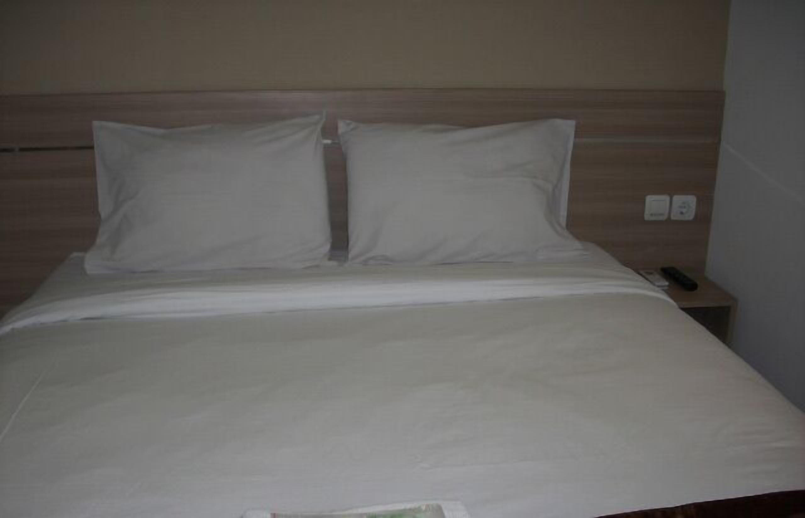 A neatly made bed with white bedding and two pillows, a wooden headboard, and a bedside table with a remote control.