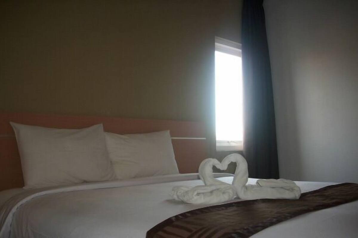 A hotel room with a neatly made bed, a heart-shaped towel arrangement, and a window with curtains.