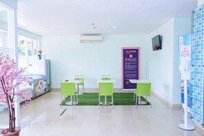 Un área de recepción con piso de césped artificial verde, mesas y sillas blancas, un cartel informativo grande y una planta en maceta.