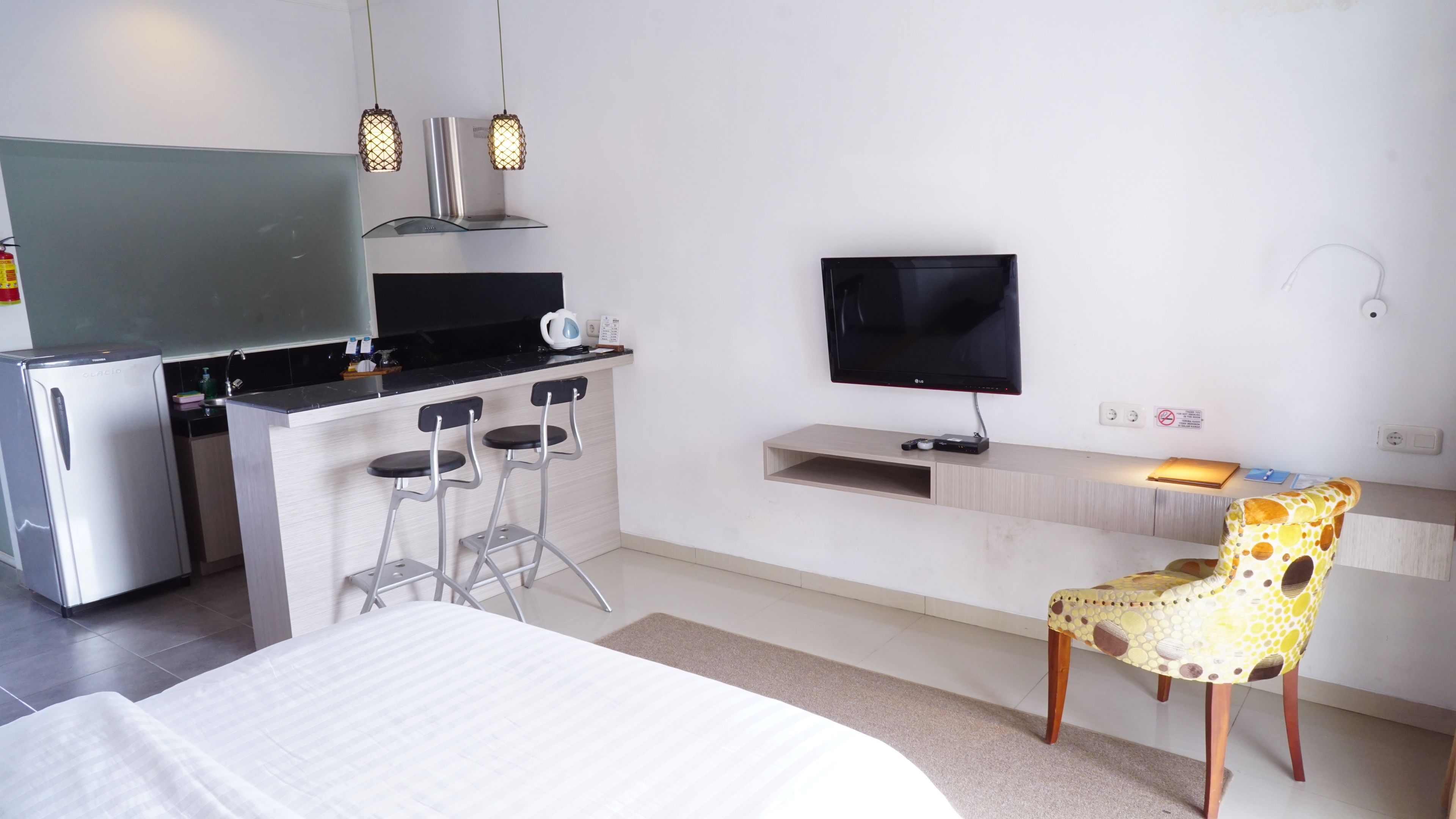 A modern hotel room with a kitchenette, a flat-screen TV, a desk with a computer, and a patterned chair.